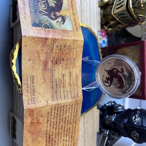 May include: A dragon-themed coin in a clear case, presented on a blue and gold decorative element. A wooden box and a brochure with text are in the background. Other decorative items, including a knight's gauntlet, are also visible.