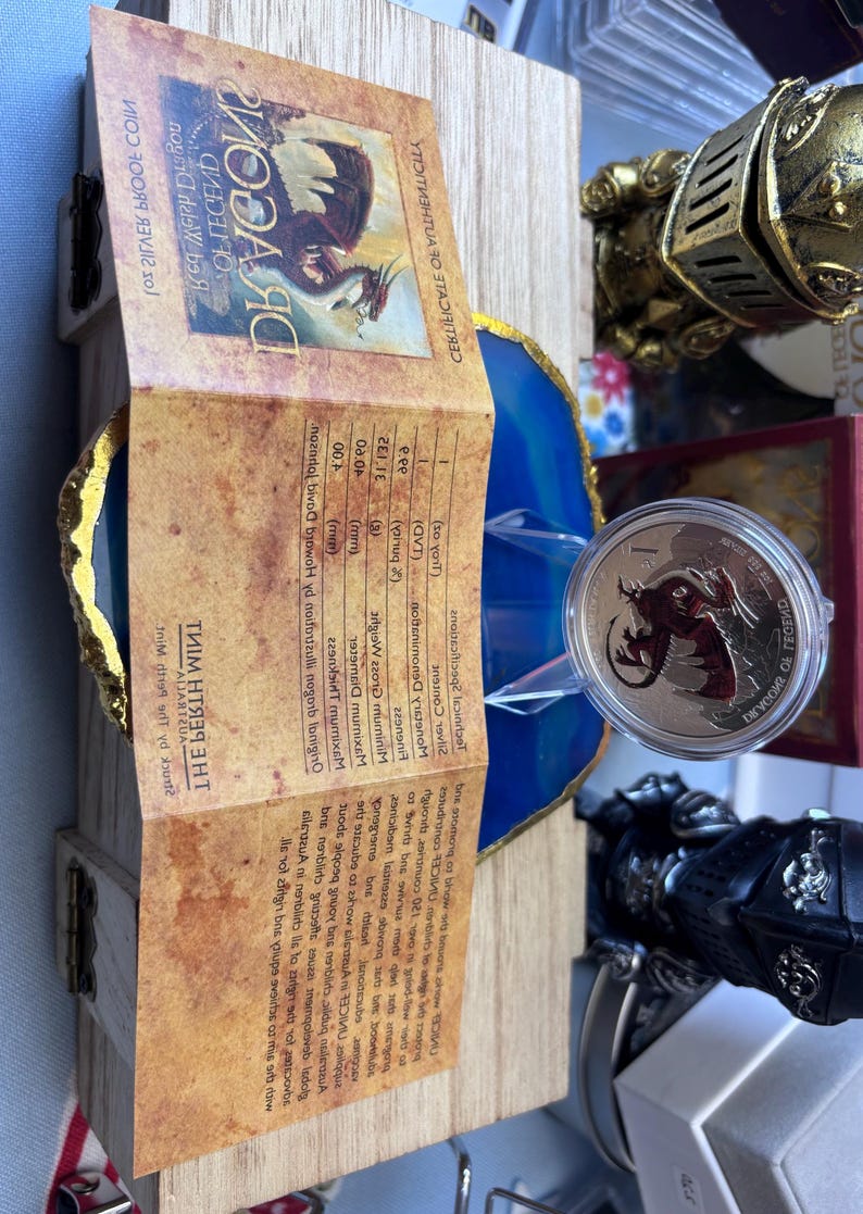 May include: A dragon-themed coin in a clear case, presented on a blue and gold decorative element. A wooden box and a brochure with text are in the background. Other decorative items, including a knight's gauntlet, are also visible.
