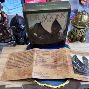 May include: A boxed silver proof coin from the "Dragons of Legend" series, featuring a European Green Dragon design. The box is green with gold lettering. Accompanying the coin is a certificate of authenticity and a wooden display stand. Decorative knight figurines are in the background.