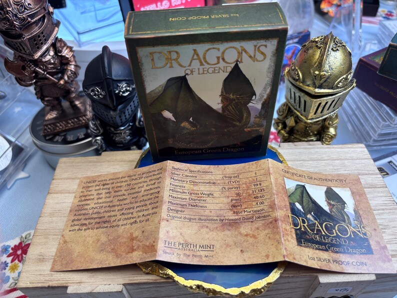 May include: A boxed silver proof coin from the "Dragons of Legend" series, featuring a European Green Dragon design. The box is green with gold lettering. Accompanying the coin is a certificate of authenticity and a wooden display stand. Decorative knight figurines are in the background.