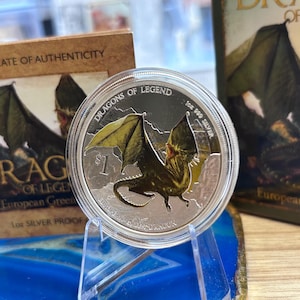 May include: A silver coin with a detailed dragon design, part of the "Dragons of Legend" series. The coin is displayed on a clear stand, with a certificate of authenticity and a book visible in the background. The coin is marked "1 oz 999 Silver".