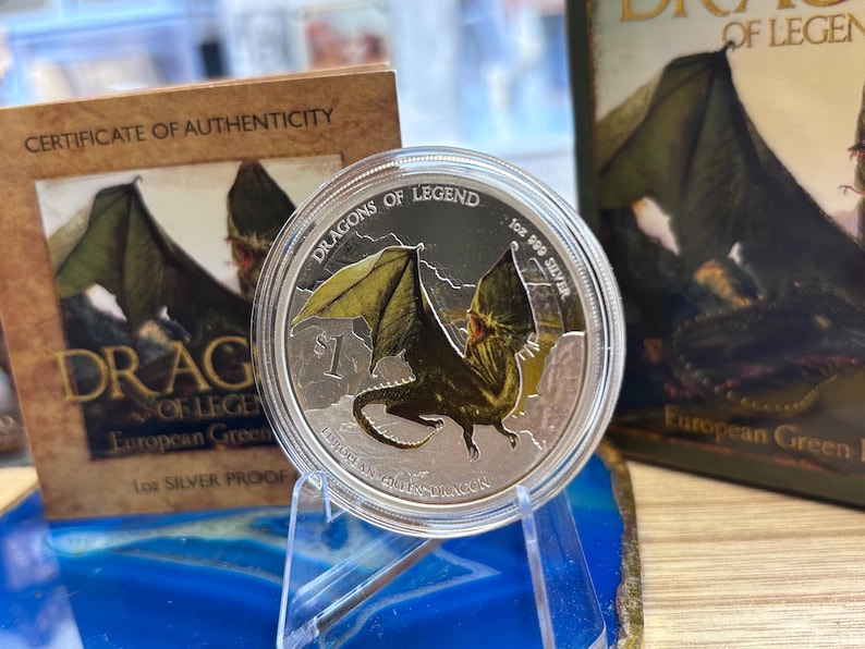 May include: A silver coin with a detailed dragon design, part of the "Dragons of Legend" series. The coin is displayed on a clear stand, with a certificate of authenticity and a book visible in the background. The coin is marked "1 oz 999 Silver".