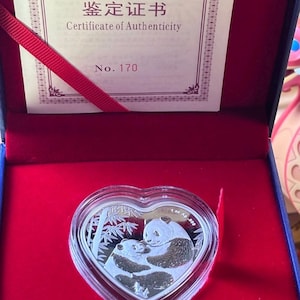 Puede incluir: Moneda de plata en forma de corazón con un panda y su cachorro, en una caja de terciopelo rojo. La moneda está marcada "1 oz Ag .999". Se ve un certificado de autenticidad, con el texto "Certificate of Authenticity" y "No. 170".
