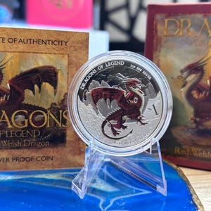 May include: A silver coin depicting a red dragon with the text "DRAGONS OF LEGEND." The coin is displayed on a clear stand, with a certificate of authenticity and a book with the same dragon design in the background. The coin is 1 oz silver.
