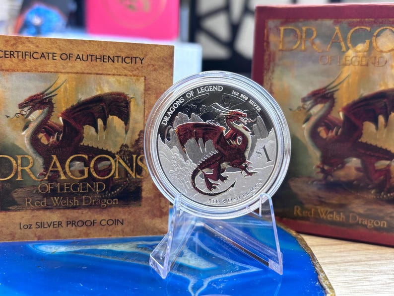 May include: A silver coin depicting a red dragon with the text "DRAGONS OF LEGEND." The coin is displayed on a clear stand, with a certificate of authenticity and a book with the same dragon design in the background. The coin is 1 oz silver.