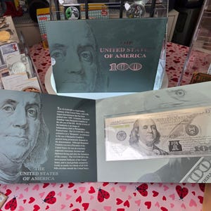 May include: A commemorative display folder featuring a United States 100 dollar bill encased in a clear protective sleeve. The folder displays a large portrait of Benjamin Franklin, the text The United States of America, and historical information.