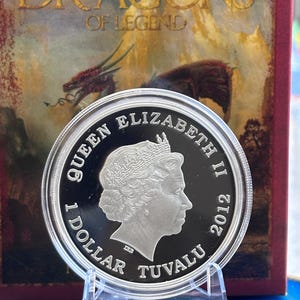 May include: A silver coin featuring Queen Elizabeth II, dated 2012, with the text "1 DOLLAR TUVALU." The coin is displayed in front of a box with the words "DRAGONS OF LEGEND" and a dragon illustration.