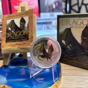 May include: A silver proof coin featuring a green dragon with outstretched wings, displayed on a clear stand. The coin is part of the "Dragons of Legend" series, with accompanying packaging and a certificate of authenticity.