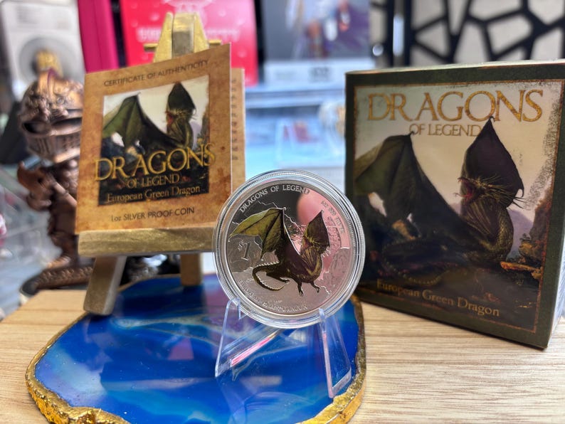 May include: A silver proof coin featuring a green dragon with outstretched wings, displayed on a clear stand. The coin is part of the "Dragons of Legend" series, with accompanying packaging and a certificate of authenticity.