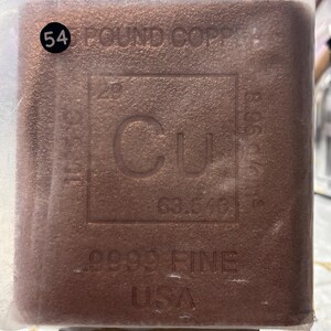 May include: A rectangular copper bar encased in a clear plastic sleeve. The surface features the chemical symbol Cu, the atomic number 29, and the atomic weight 63.546. Text includes 54 POUND COPPER, 9999 FINE, and USA. The bar has a textured, metallic finish.