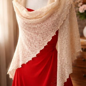 Puede incluir: Un chal de punto color crema con un delicado encaje está drapeado sobre un vestido rojo. El chal está envuelto alrededor de los hombros y tiene un borde festoneado. El vestido es de color rojo vibrante.