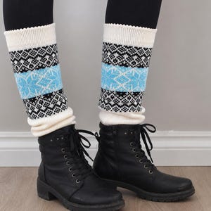 May include: A pair of knit leg warmers featuring a cream-colored base with a horizontal band of light blue and black geometric snowflake patterns. The leg warmers have a ribbed texture at the top and bottom edges and are worn over black boots.