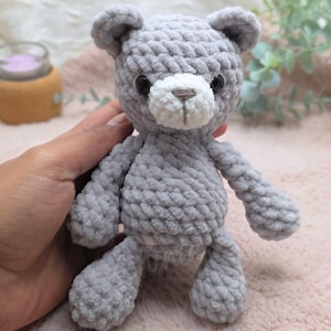 Könnte beinhalten: Ein handgehäkelter Teddybär in Grau mit weißer Schnauze und schwarzen Augen. Der Bär hat eine weiche, plüschige Textur. Es ist ein niedliches und kuscheliges Spielzeug, ideal für ein Kind oder als Dekorationsartikel.