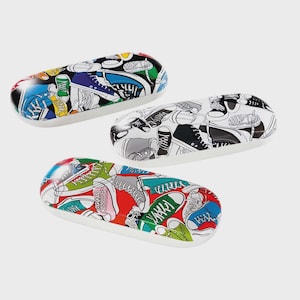 May include: Three oval-shaped spectacle cases with a white base and a colourful sneaker pattern. The cases feature a variety of shoe designs in black, white, red, green, blue, and yellow. The cases are arranged on a light grey background.