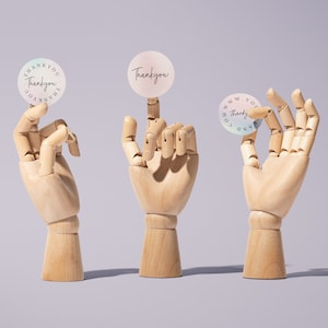 Pu&ograve; includere: Tre mani di legno che tengono adesivi rotondi con il testo "Thank you" e "www.andco.com Thanky" in scrittura nera su uno sfondo blu chiaro e rosa.