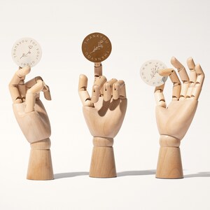 Pu&ograve; includere: Tre adesivi rotondi con disegni floreali e le parole "grazie per il tuo ordine" e "www.yourdomain.com" sono mostrati da mani di legno. Un adesivo &egrave; marrone e due sono bianco sporco.