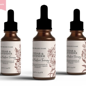 Op de afbeelding: Drie amberkleurige glazen flessen met druppelaars en witte etiketten. De etiketten bevatten een bloemenmotief en de tekst "Cedar & Lavender Essential Moisture Therapy", "geperste essenti&euml;le olie", "Gemaakt van biologische producten" en "340G e 13.4OZ".