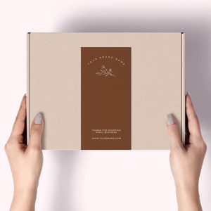 Pu&ograve; includere: Una scatola di cartone beige con un'etichetta rettangolare marrone. L'etichetta presenta il testo "YOUR BRAND NAME" e "THANKS FOR SHOPPING SMALL BUSINESS" con un design floreale. &Egrave; visibile anche l'indirizzo del sito web.