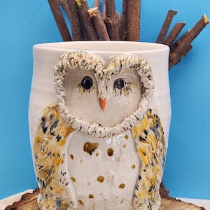 May include: A white ceramic owl-themed mug with a textured owl face and wings. The owl's face has black eyes and an orange beak. The wings are decorated with yellow, blue, and black patterns. The mug is on a wooden surface.
