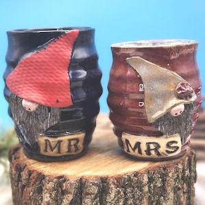 May include: Two ceramic gnome-themed mugs, one navy blue with a red hat and the word "MR" and the other burgundy with a silver hat and the word "MRS". The mugs are on a wooden surface.