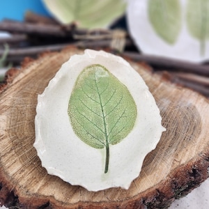 May include: A small, white ceramic dish with a green leaf imprint. The leaf's veins are visible, and the dish has a textured, organic edge. The dish is displayed on a wooden surface, with other similar dishes in the background.