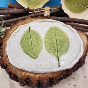 May include: A handmade ceramic dish with two green leaf imprints on a white background, set on a rustic wooden slice. Additional leaf-imprinted ceramics and wooden sticks are visible in the background, suggesting a nature-inspired theme.