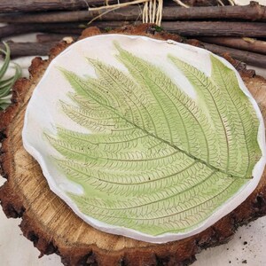 May include: A handmade ceramic dish with a fern leaf design. The dish is white with a green fern leaf imprint. It sits on a rustic wooden slice, with a natural, organic aesthetic. The dish is approximately 6 inches in diameter.