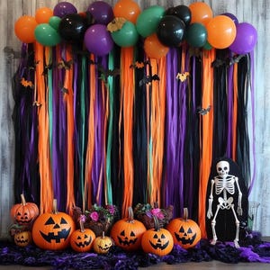 May include: A Halloween party backdrop featuring a balloon arch in orange, purple, green, and black, with hanging streamers in matching colors, paper bats, carved jack-o'-lantern pumpkins, and a standing skeleton figure on a purple fabric base.