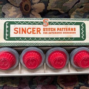 Puede incluir: Un juego de patrones de puntada Singer vintage para una zigzagueadora automática. La caja es blanca con ribete verde y el logotipo de Singer. Se ven cuatro selectores de patrones rojos y grises dentro de la caja.