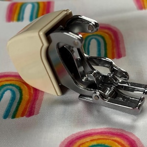 Könnte beinhalten: Ein Nähmaschinen-Nähfuß mit metallisch silberner Oberfläche und einem cremefarbenen Kunststoff-Schaftansatz. Der Mechanismus verfügt über ein bewegliches Vorderteil mit zwei parallelen Zinken zur Stoffführung beim Nähen mit der Maschine.