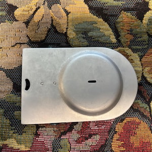 Puede incluir: Un objeto metálico de color plateado con una base rectangular y una parte superior redondeada. Presenta una hendidura circular y una pequeña ranura horizontal. También son visibles dos pequeños agujeros y un recorte en forma de media luna.