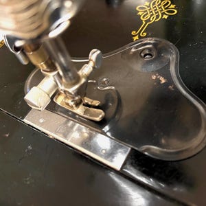 May include: A black vintage sewing machine featuring a metallic needle bar and presser foot assembly. The machine base is decorated with gold-colored filigree patterns and includes a black plastic throat plate cover with a smooth, contoured shape.