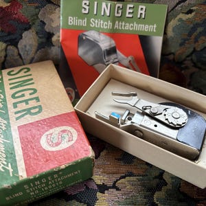 May include: A vintage Singer Blind Stitch Attachment in a cream-colored box with a matching instruction manual. The metal sewing machine accessory features a black side panel, a silver gear-like dial, and a small blue plastic component.