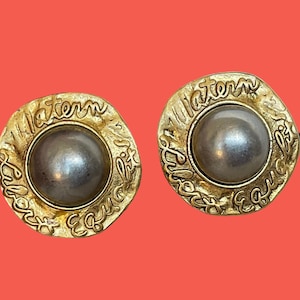 May include: A pair of gold-tone earrings with a pearl-like center and the words "maternity" and "equality" inscribed around the edge.