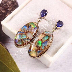 May include: A pair of silver earrings featuring oval-shaped opal pendants with a mosaic of colors, including blue, green, and gold. Each earring has a small, teardrop-shaped, dark blue gemstone at the top.
