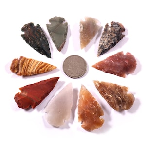 以下が含まれることがあります： 10個のカラフルな手作りの石の矢じりのコレクション。硬貨を中心に円形に配置されています。矢じりは赤、茶色、ベージュ、灰色など様々な色があり、白い背景に表示されています。