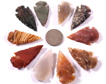 May include: A collection of ten colorful, handcrafted stone arrowheads arranged in a circle around a quarter. The arrowheads vary in color, including red, brown, tan, and gray, and are displayed on a white background.