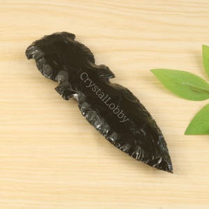 May include: A black obsidian arrowhead with a textured surface. The arrowhead is on a light wood surface. The text "CrystalLobby" is printed on the arrowhead.