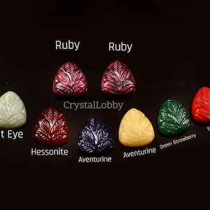 May include: Assortment of faceted gemstone cabochons in various colors, including ruby, cat eye, hessonite, aventurine, green strawberry, and synthetic. The cabochons are shaped like leaves and are arranged in a row on a black background. The text "CrystalLobby" is visible in the center of the image.