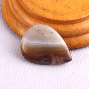 May include: A teardrop-shaped gemstone with a polished surface. The stone features bands of white, tan, and brown hues, with darker speckling near the base. The gem is set against a light background and a wooden base.
