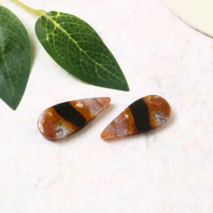 Peut inclure: Deux perles en forme de goutte de pierres précieuses. Les perles présentent un mélange de couleurs, notamment du marron, de l'orange et du blanc, avec une bande noire proéminente au centre. Les perles sont lisses et polies, avec une finition brillante.