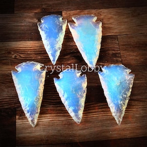 May include: Five opalescent arrowhead-shaped crystals with a blue and white iridescent glow. The crystals are arranged on a dark wood surface. The text "crystalLobby" is visible on the surface.
