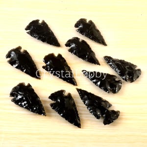 May include: Ten black obsidian arrowheads, each with a pointed tip and a slightly curved base, arranged on a light wood surface.