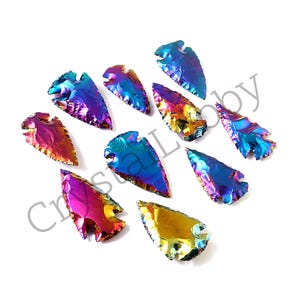 Può includere: Una collezione di ciondoli a forma di punta di freccia in vetro iridescente. I ciondoli mostrano uno spettro di colori, tra cui blu, viola e oro. Ogni punta di freccia ha una punta appuntita e una base dentellata. I ciondoli sono disposti su sfondo bianco.