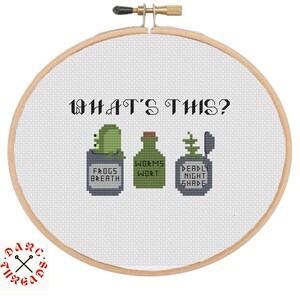 May include: A cross-stitch pattern in a wooden embroidery hoop featuring the text WHAT'S THIS? above three potion bottles labeled FROGS BREATH, WORMS WORT, and DEADLY NIGHT SHADE. The design uses green, grey, and black thread on white fabric.
