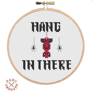 Pode incluir: Um aro de bordado branco com um desenho de ponto cruz preto e vermelho. O desenho diz "Hang in there" com uma aranha vermelha e preta pendurada na palavra "Hang".