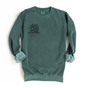 May include: A teal-colored sweatshirt with a black graphic of a dog's face and the name "Bentley" printed below. The sweatshirt has a crew neck and long sleeves with cuffed wrists. The fabric appears soft and comfortable.