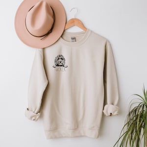 May include: A beige sweatshirt with a black and white illustration of a dog peeking out of a pocket. The dog's name, "Tucker", is written below the illustration.