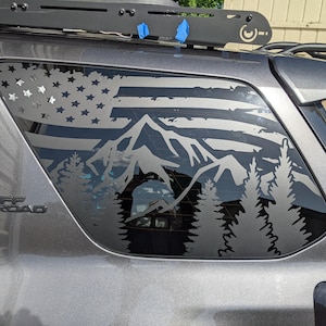 May include: A gray vehicle window decal featuring a mountain range and a stylized American flag design. The decal includes stars, stripes, and silhouettes of trees. The vehicle has a roof rack and the text "TRD OFF ROAD".