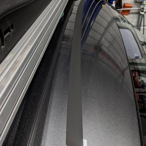 May include: A close-up shot of a gray car roof with a black strip running down the center. The roof has a metallic finish, and a roof rack is visible on the left side. The background shows a garage setting.